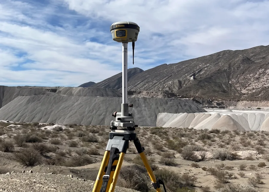 Receptor GNSS sobre trípode, con acopios de material al fondo.